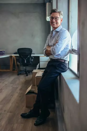 Male entrepreneur standing in office with his arms crossed. Businessman leaning to a window sill at his office.